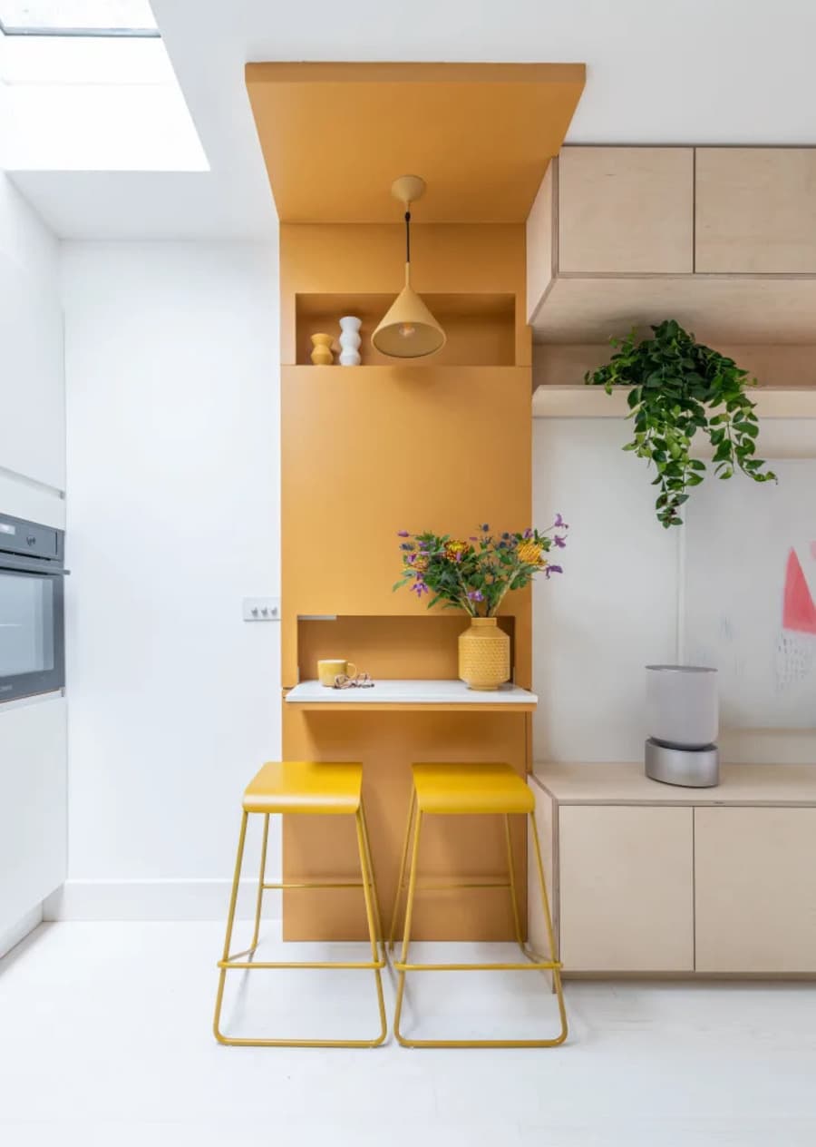 Cocina pequeña con muros y  piso blanco. Tiene el sector del muro pintado amarillo, con una mesa plegable y dos pisos amarillos. Muebles de cocina de madera color crema o beige. Repisa flotante con una planta. Horno empotrado