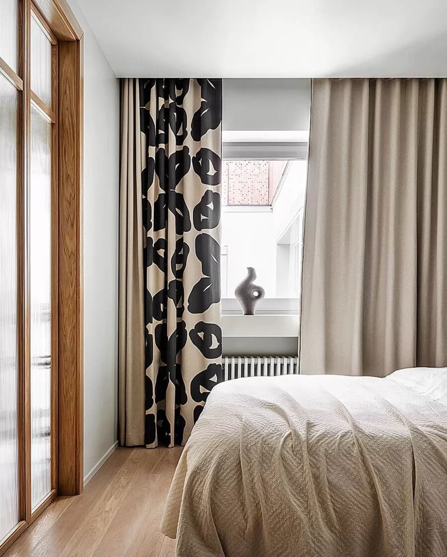 Dormitorio moderno con muros blancos y gran ventana con cortinas combinadas, color beige y cortinas estampadas con figuras negras. Cama con cubrecama color beige.