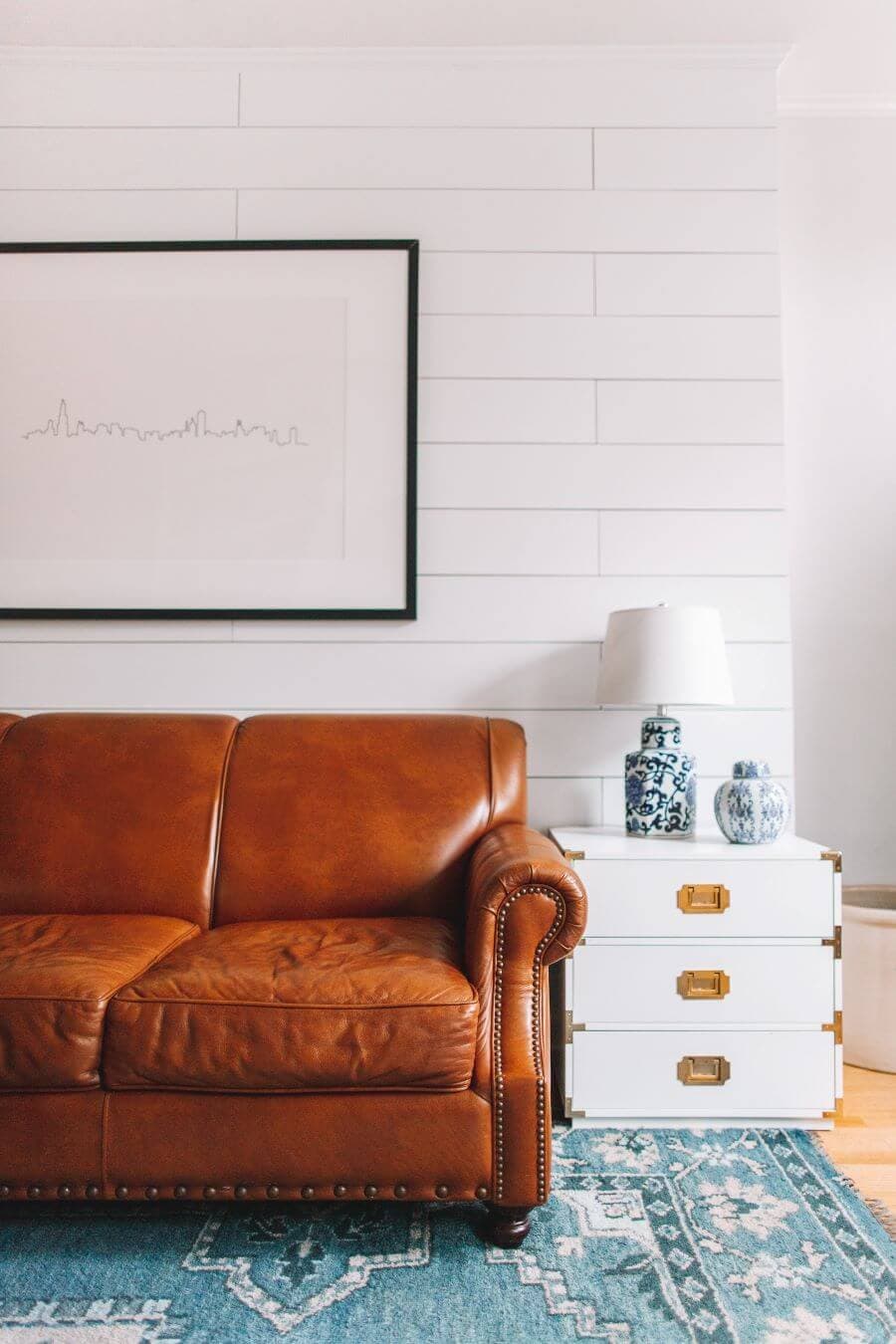 Living estilo nórdico minimalista, con muro de madera blanca. Sillón de cuero café, junto a él una mesa blanca con tres cajones con tiradores dorados. Sobre el sillón, en la pared hay un cuadro de un skyline hecho con una línea. Lámpara de mesa de cerámica