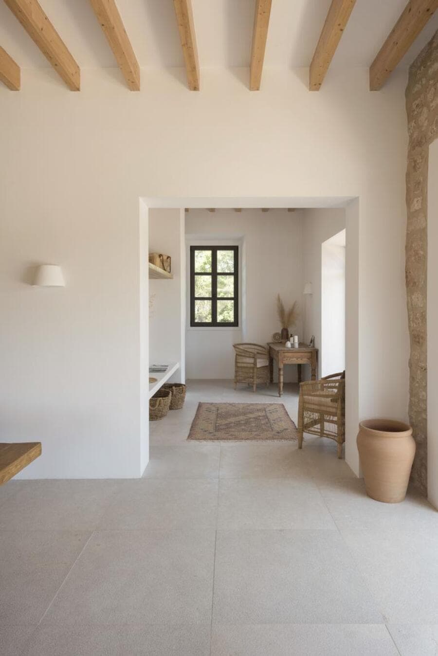 Casa estilo mediterráneo, minimalista. Muros blancos con piso de cerámica beige. Dos sillas de mimbre, una mesa cuadrada de madera en un rincón. Al centro hay una alfombra estilo oriental en tonos tierra. En la esquina un gran jarrón de cerámica color café
