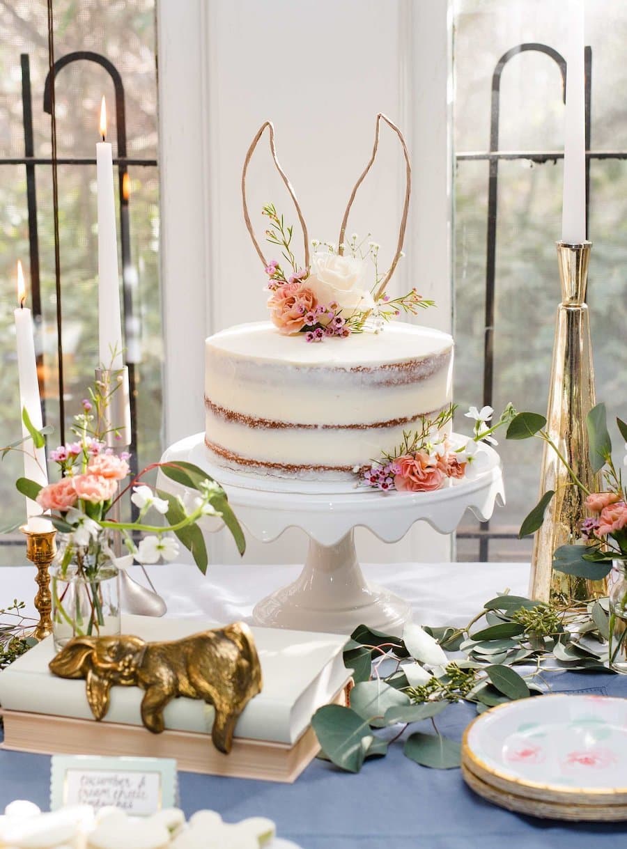 Detalle de una mesa de celebración de baby shower con temática de cuento, hay hojas decorativas, candelabros dorados, flores rosa pastel, un conejo dorado y una torta blanca con flores y orejas de conejo.