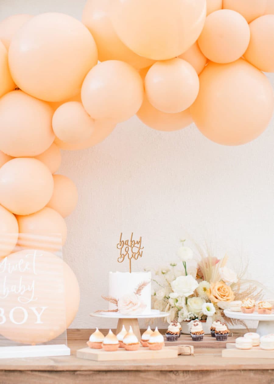 Mesa con comida para un baby shower con flores y cupcakes en tonos anaranjados y amarillos, una columna de globos de distintos tamaños de color salmón y una torta blanca.