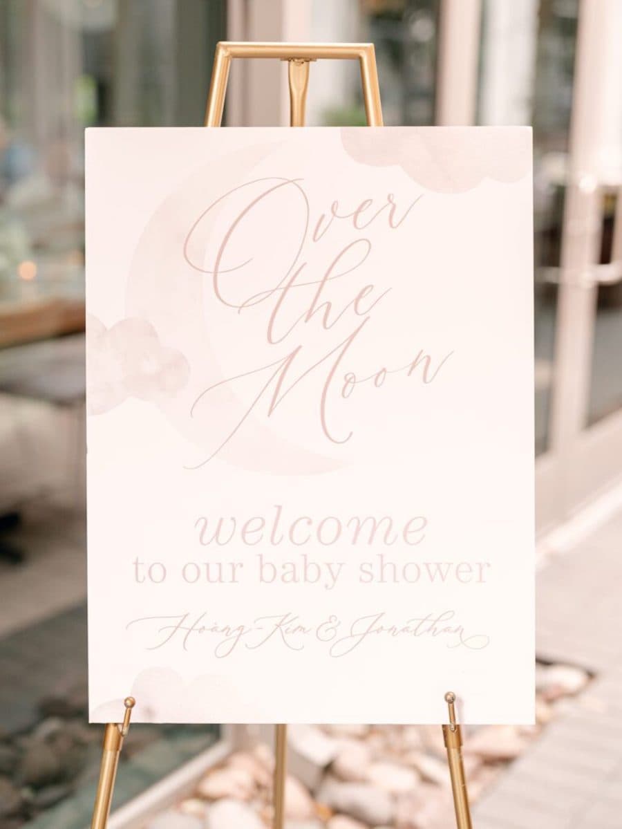 Letrero de bienvenida para un baby shower con diseño de nubes y luna en tonos rosa pastel.