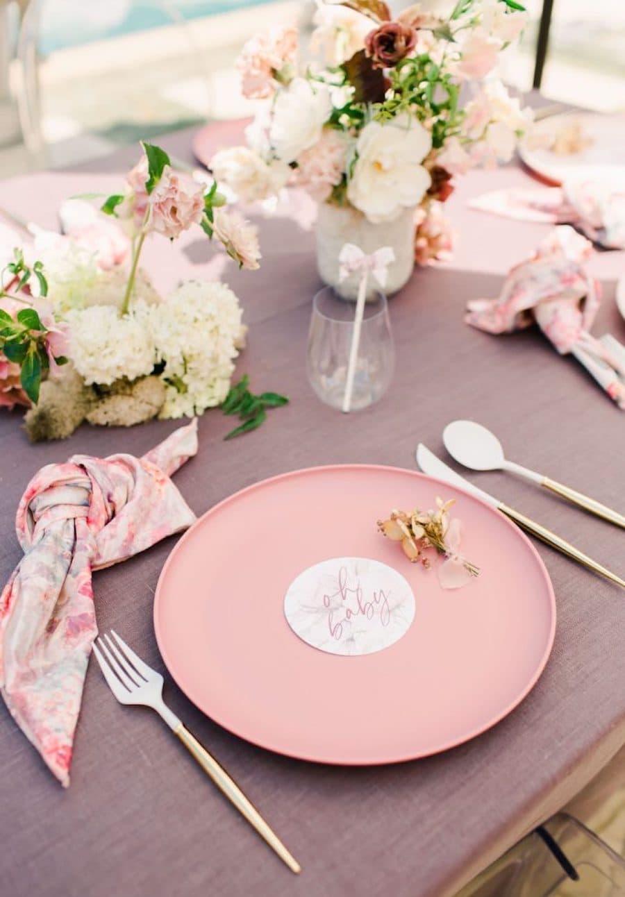 Detalle de una mesa con mantel morado, un plato rosado, cubiertos plateados, servilletas rosadas y centro de mesa con flores blancas.