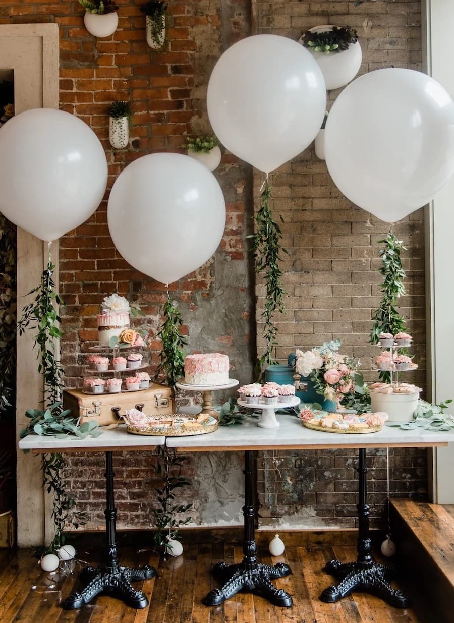 Mesa de baby shower con comida sobre ella, hay cupcakes, una torta y otros dulces, así como 4 globos blancos con ramas que sirven como decoración.