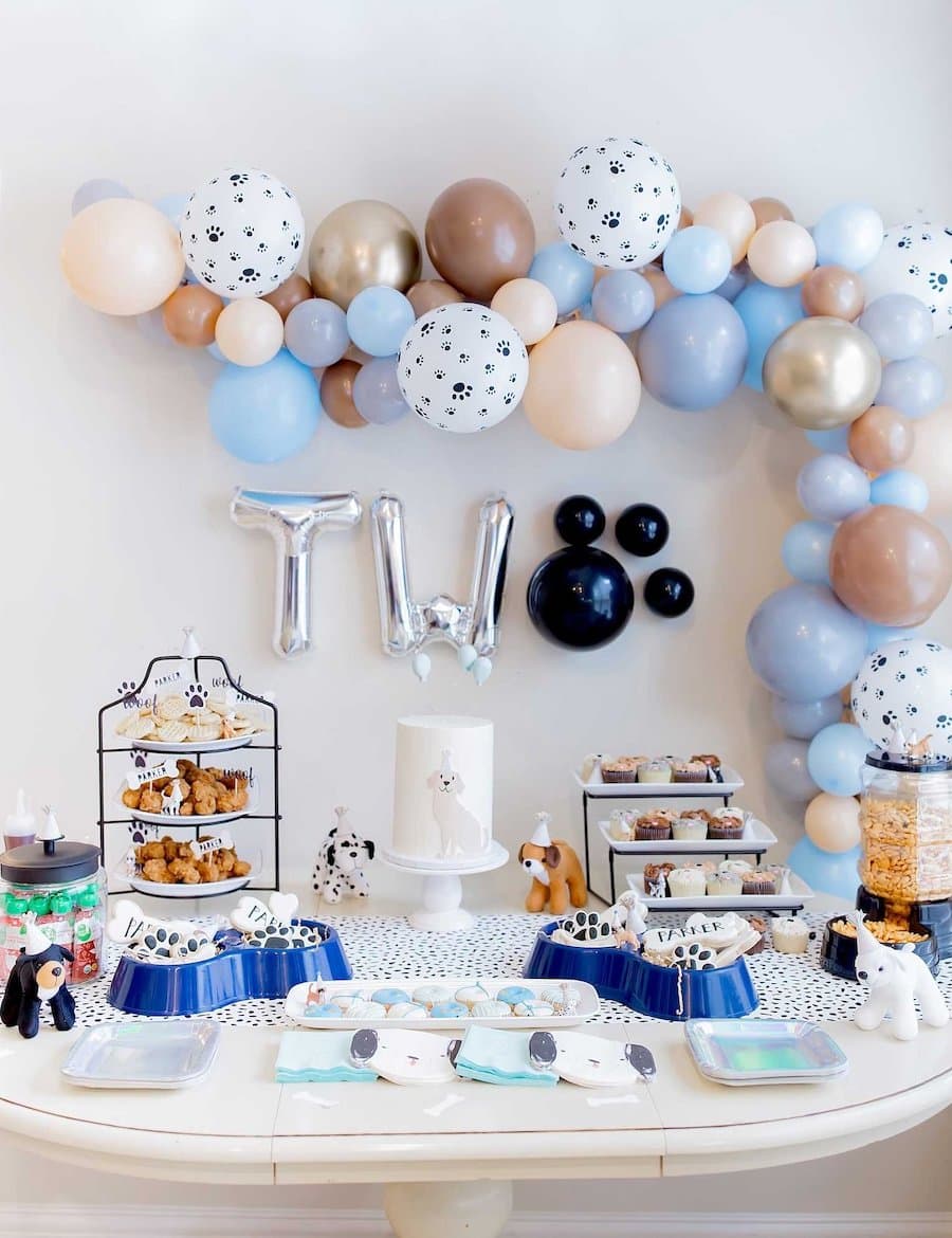 Mesa de cumpleaños con dulces, cupcakes, pasteles, una torta y decoraciones con motivos de perros. En la pared hay una especie de arco con globos de colores de diferentes tamaños.