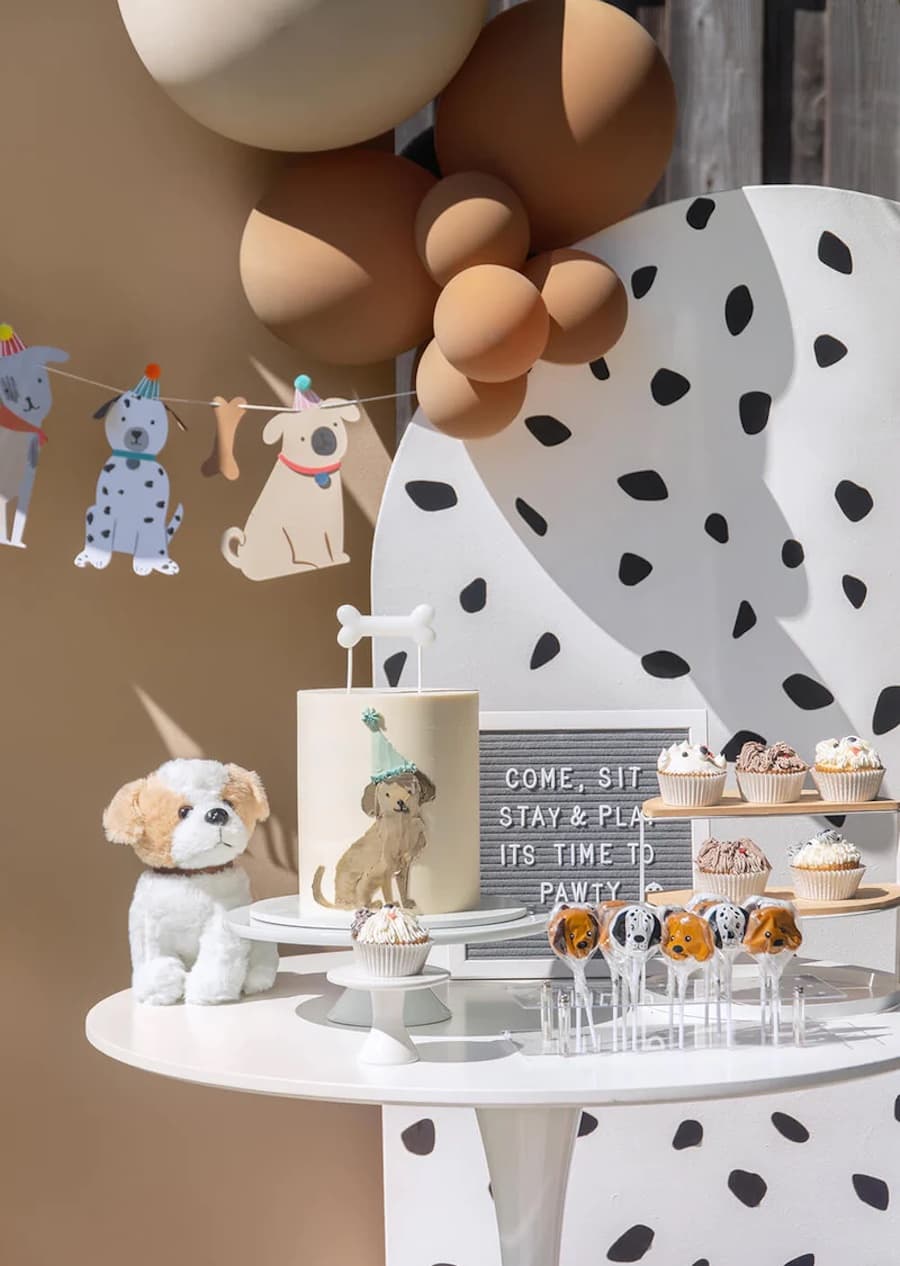 Mesa de cumpleaños con motivos de perros. Hay una torta, un peluche, cupcakes y pasteles. Detrás hay globos y guirnaldas con dibujos de perros.