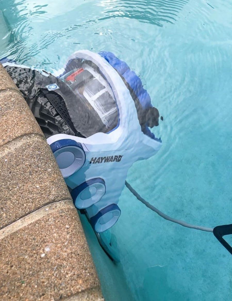 Aspiradora robot para piscina, sumergida en el agua, subiendo por una de las paredes. El borde de la piscina es de pequeñas piedras color café.