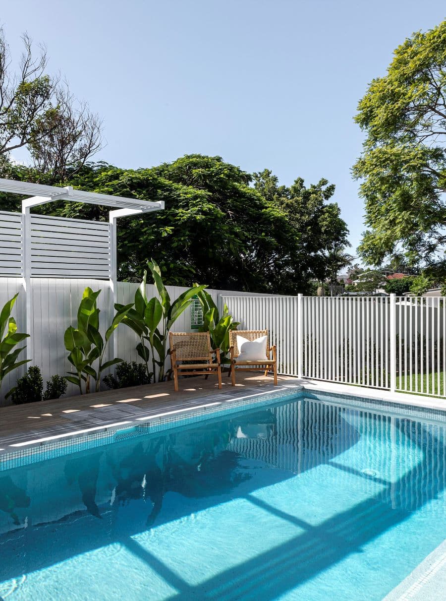 Gran piscina en un patio. Junto a ella hay un deck de madera clara con dos sitiales o reposeras de madera y mimbre, una de ellas con un cojín blanco. La piscina está rodeada por una reja de seguridad blanca y por plantas tropicales.