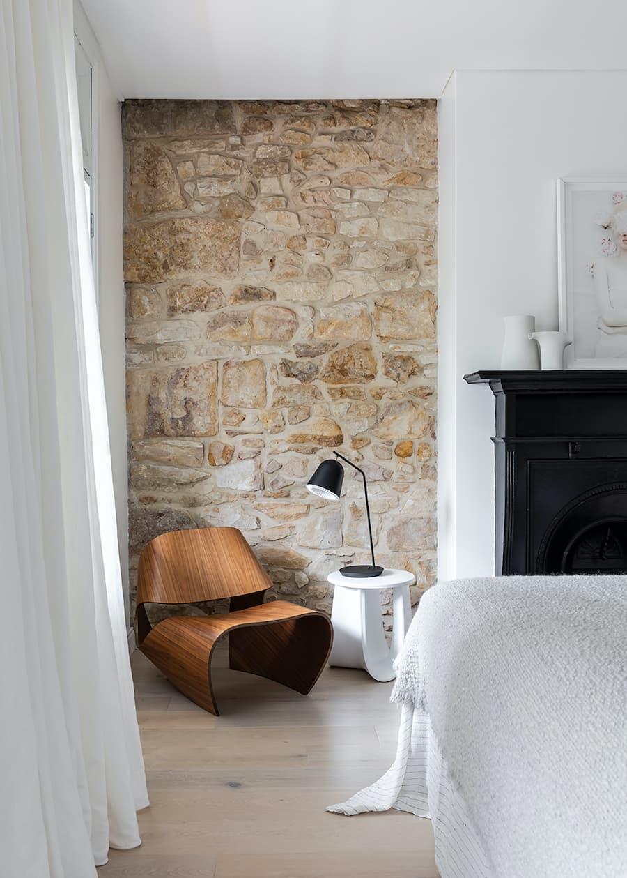 Rincón de una sala de estar con un muro de acento de piedra en tonos arena y anaranjados. Junto a él hay un sillón de diseño de madera, una mesa lateral blanca y sobre ella, una lámpara de mesa negra. 