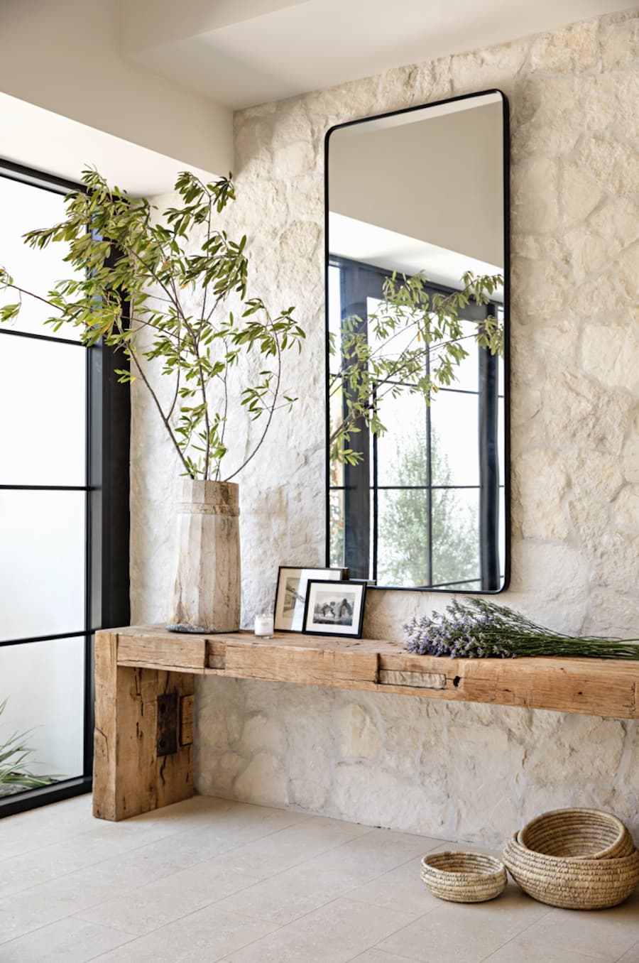 Entrada de una casa con un muro de piedra con un arrimo de madera rústica. Sobre él hay una planta, cuadros con fotos y flores. En el muro hay un espejo rectangular con marco negro.