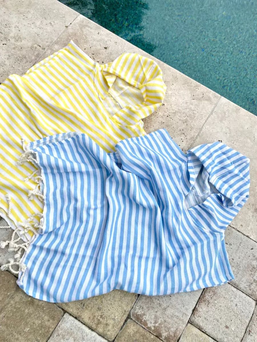 Dos toallas de playa, tipo poncho, sobre el suelo junto a una piscina. Una es blanca con líneas amarillas y la otra es blanca con líneas azules.