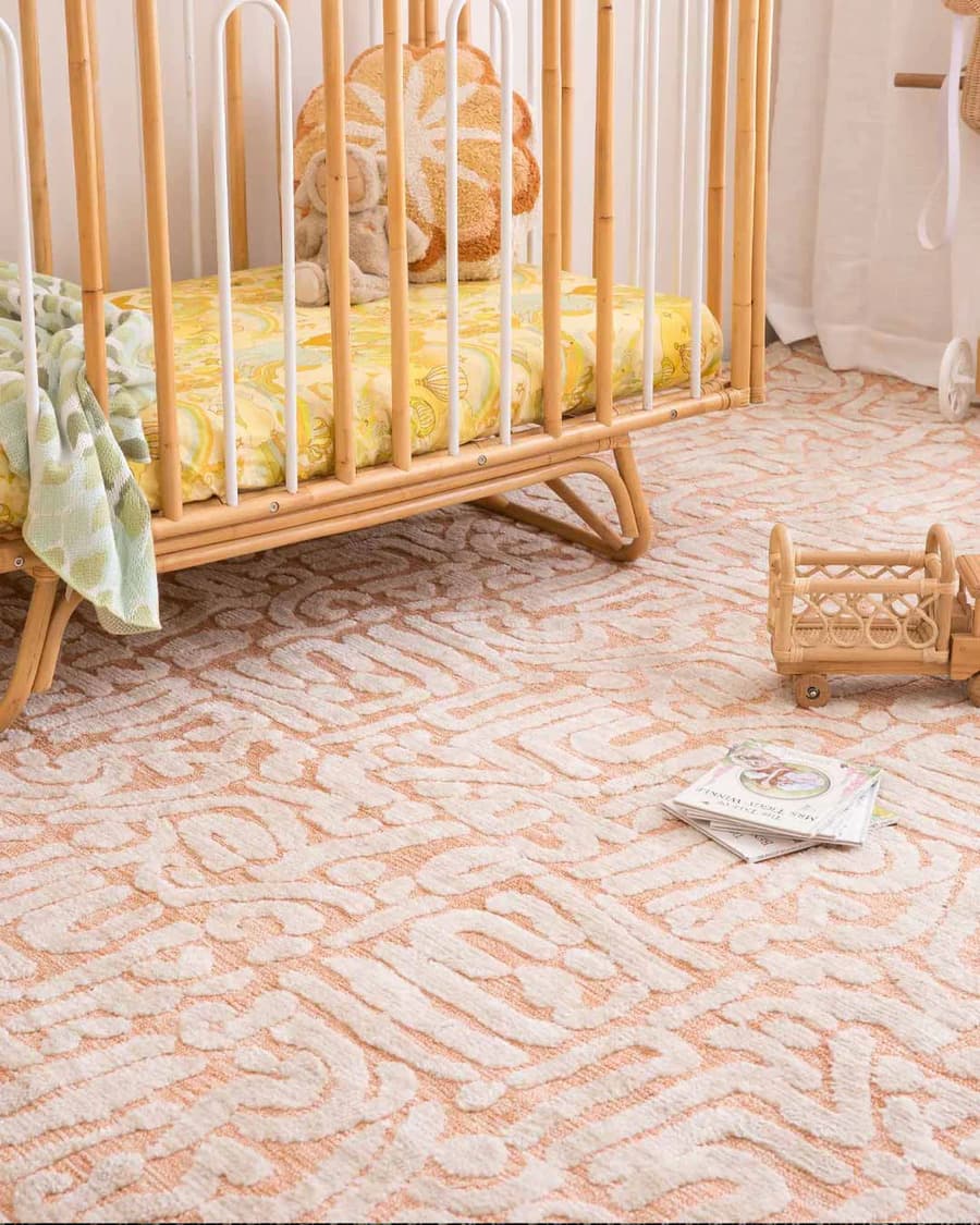 Dormitorio infantil con alfombra con patrones de color Peach fuzz. Cuna de madera con cubre cama amarillo, manda verde, un cojín con forma de flor y un muñeco. En el suelo hay un camión de madera y mimbre y libros.