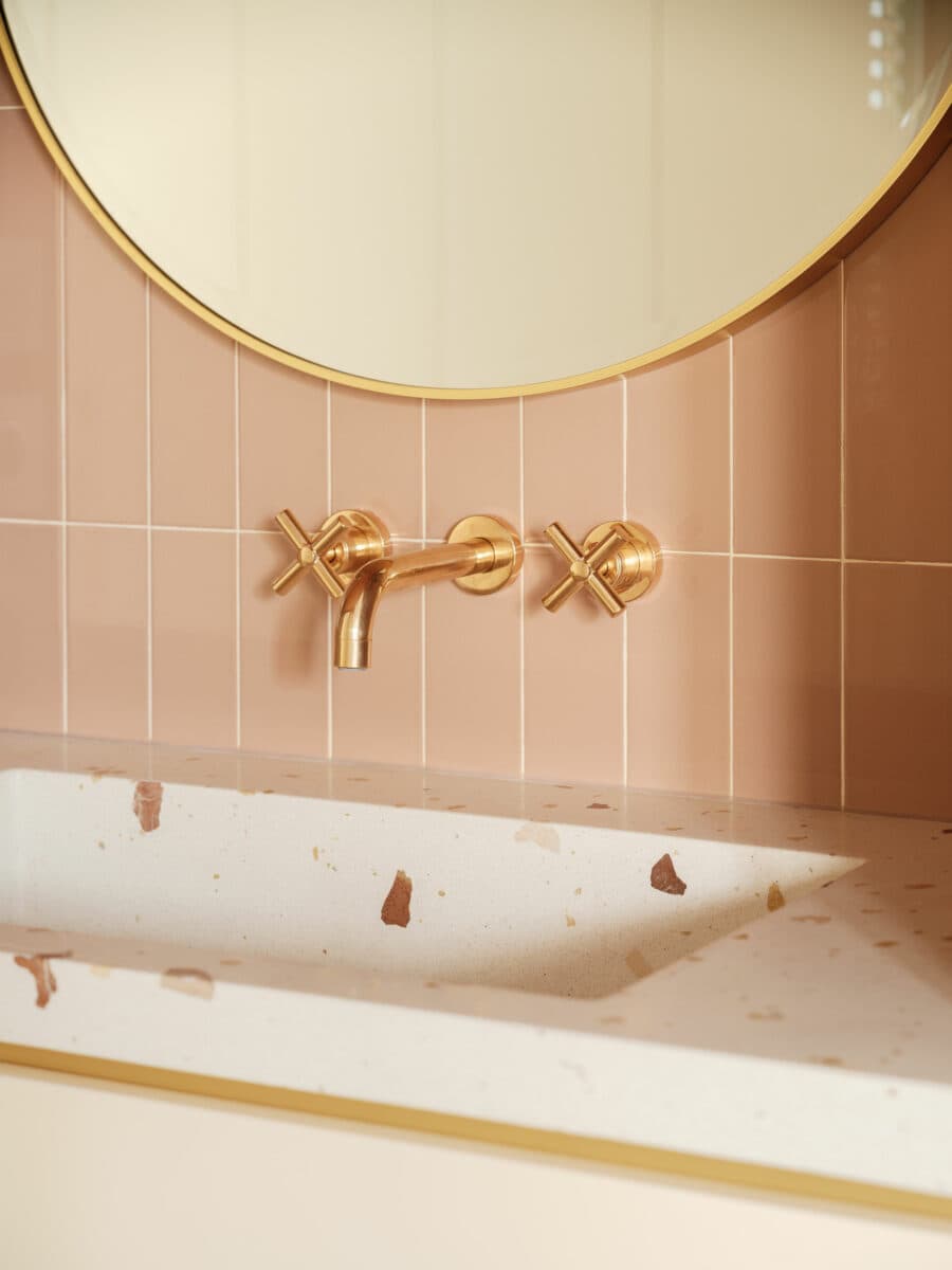 Detalle de un lavamanos de un baño estilo mid century. Superficie de terrazo, llaves al muro doradas, cerámicas alargadas color Peach fuzz. Se ve la parte de un espejo redondo con marco dorado