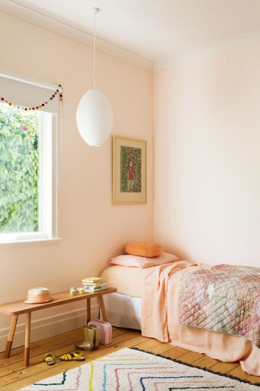 Dormitorio infantil con muros pintados de color Peach fuzz. Cama de una plaza junto a la pared, con sábanas color damasco y un cobertor. Lámpara colgante de papel, banqueta de madera bajo una ventana. Piso de madera y alfombra blanca con líneas de colores