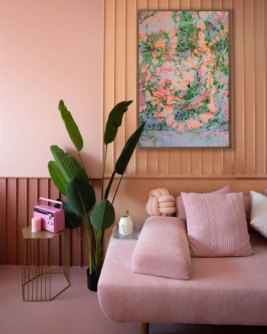 Living con parte del muro con revestimiento de madera. Las paredes tiene varios colores: rosado, Peach fuzz y rojo oscuro. Piso rosado y sofá palo rosa. Junto al sillón hay una planta tropical verde oscuro. Gran cuadro con diseño marmolado. 