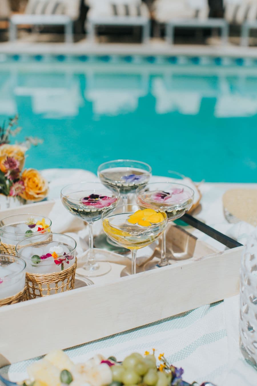 Detalle de bandeja de madera con 4 copas de martini con hielos con flores comestibles. 4 vasos altos con agua. Toso está sobre una toalla blanca y celeste. Atrás se ve una piscina con el reflejo de unas reposeras.