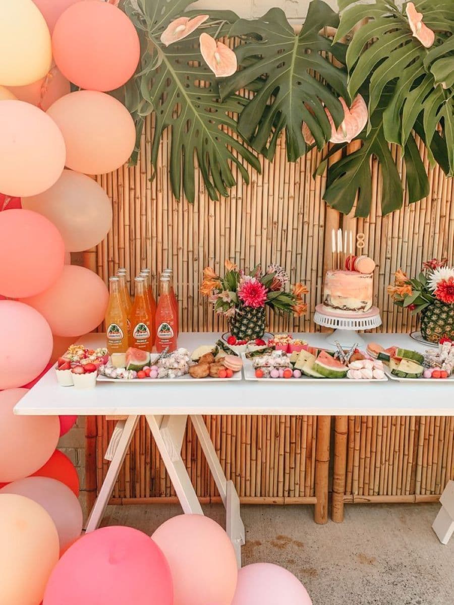 Mesa de comida en pool party. Mesa hecha con una base blanca y caballetes. Sobre ella hay dos tablas con frutas y dulces, una torta y aguas. Dos piñas con flores como decoración. Al costado hay una estructura decorativa de globos en color rosado y damasco.