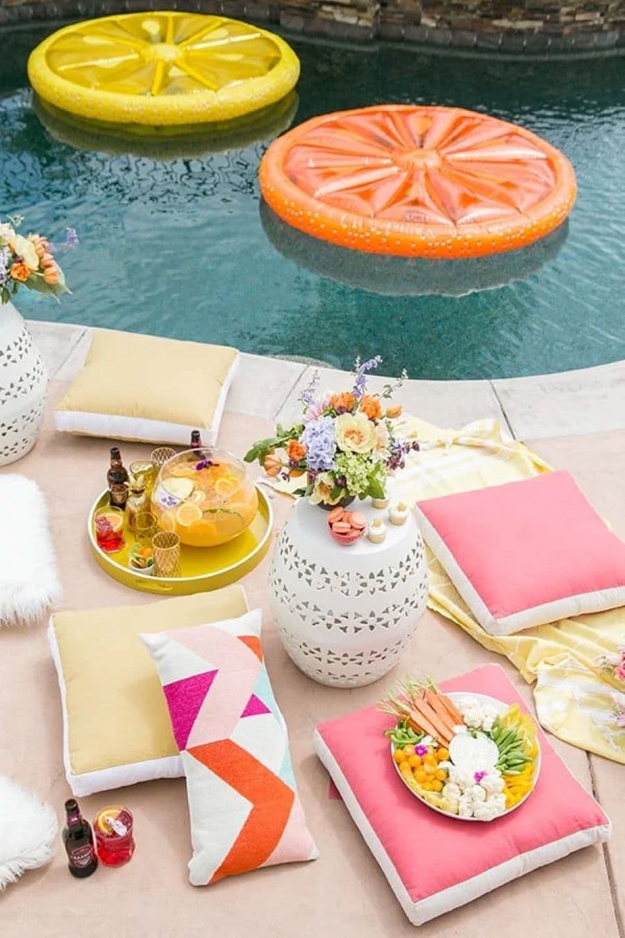 Imagen cenital del borde de una piscina, se ve el agua y una zona de descanso. Hay 4 cojines cuadrados de color amarillo y rosado, y un cojín alargado con diseño geométrico. Mesa de centro blanca con ramo de flores. Bandeja amarilla y flotador de naranja.