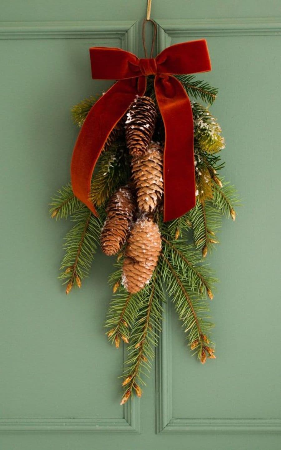 Detalle de una puerta de madera verde, con un adorno navideño hecho con ramas de pino, semillas de pino y una cinta de terciopelo roja.