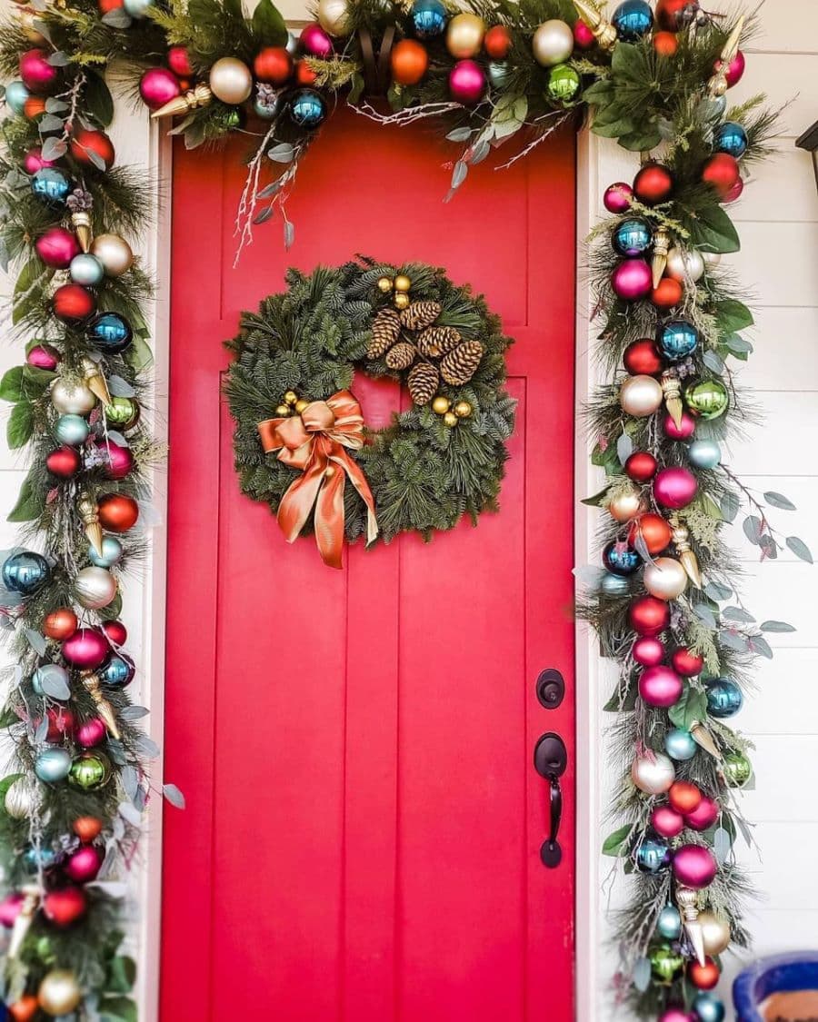 Puerta roja de entrada, con una guirnalda navideña verde, con semillas y una cinta. Alrededor de la puerta hay una guirnalda con ornamentos redondos de diferentes colores y ramas de eucaliptus.