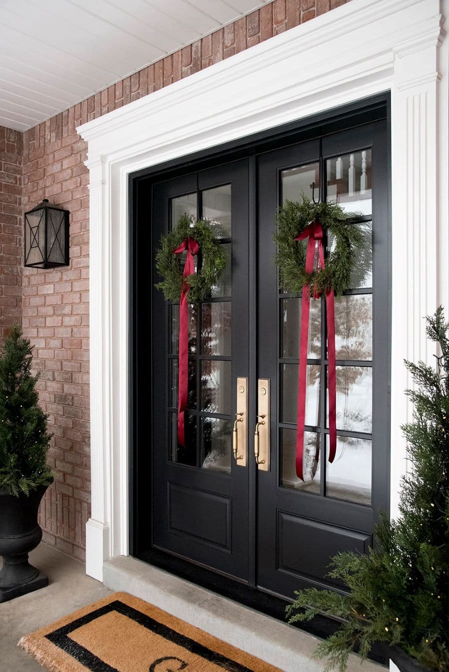 Gran puerta doble de entrada. Es de madera negra y tiene ventanas. De cada puerta cuelga una corona navideña de pino, con una cinta roja colgando. Casa con muros de ladrillo y dos pinos a los costados de la puerta.