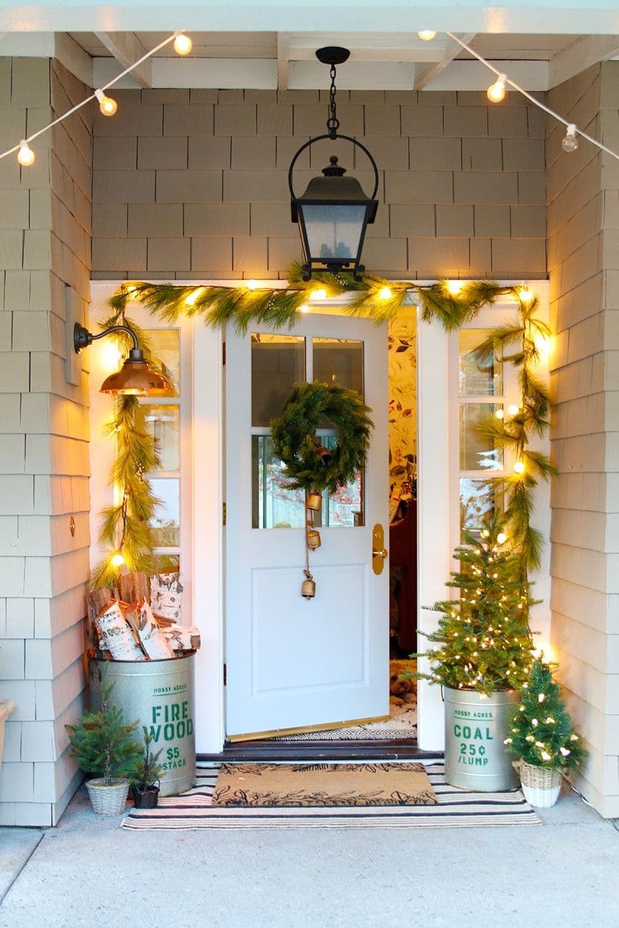 Entrada de una casa con una guirnalda de pino y luces encendidas alrededor de la puerta. La puerta es blanca y está abierta, tiene una corona navideña verde con campañas doradas colgando. Balde metálico con leña y otro balde con un pino