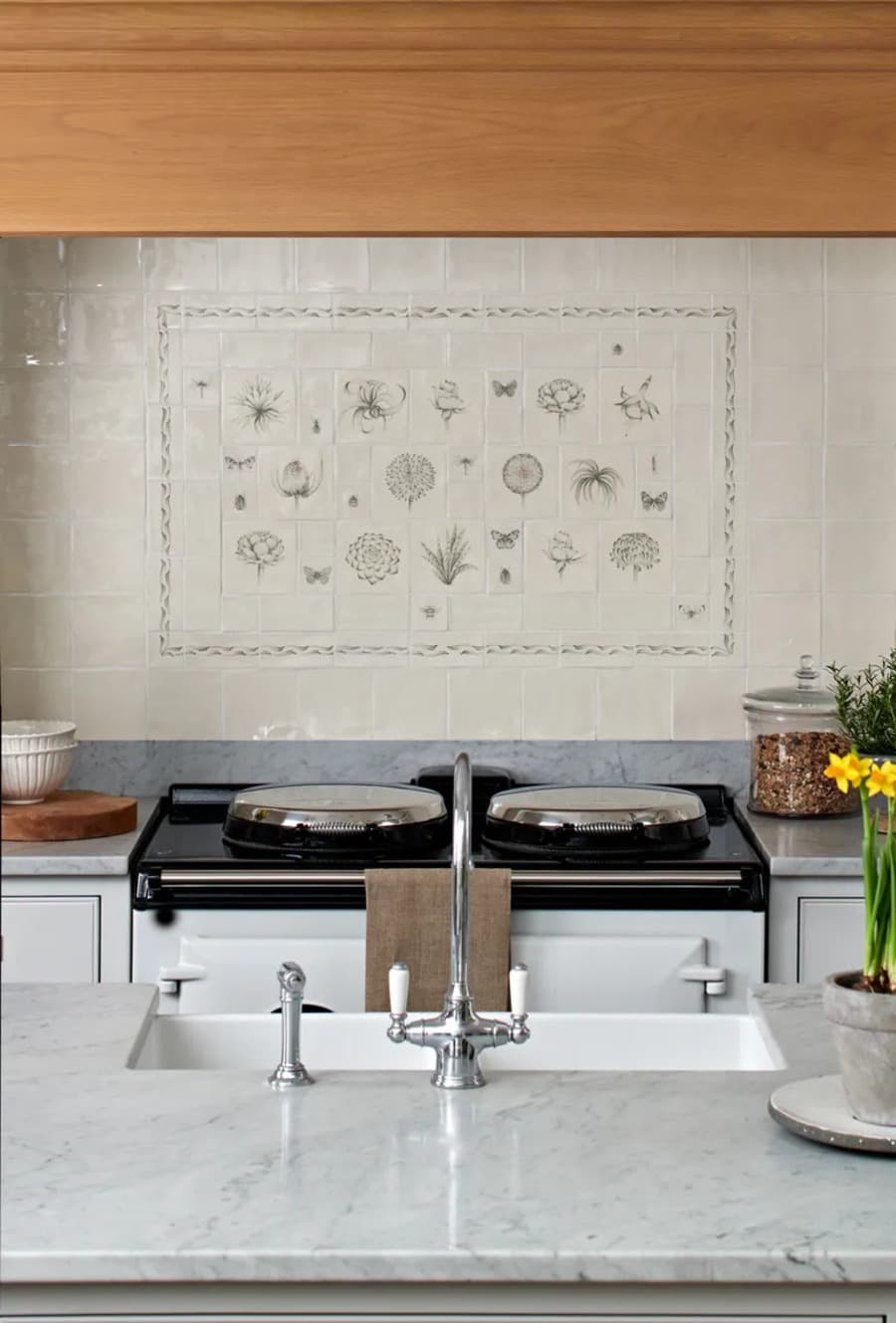 Cocina clásica con muro con azulejos color beige verdoso. Al centro hay un diseño con azulejos pintados  mano con flores, mariposas y hojas. Muebles de cocina blancos, grifería cromada estilo vintage.
