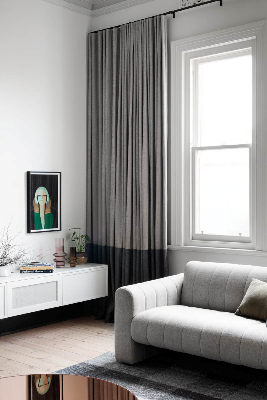 Living moderno con gran ventana con una cortina bicolor, gris claro y gris oscuro. Sillón gris de estilo contemporáneo. Junto al muro blanco hay un mueble bajo, blanco, con puertas. Sobre él hay adornos, libros y plantas. 