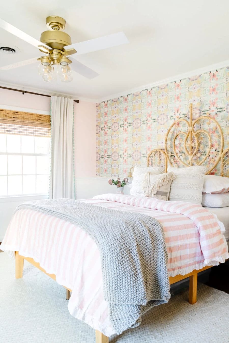 Dormitorio estilo romántico, con gran cama king con plumón a rayas rosado con blanco. Respaldo de mimbre. Ventilador de techo dorado, con luces incorporadas. Pared rosada y un muro con papel mural vintage. Alfombra gris