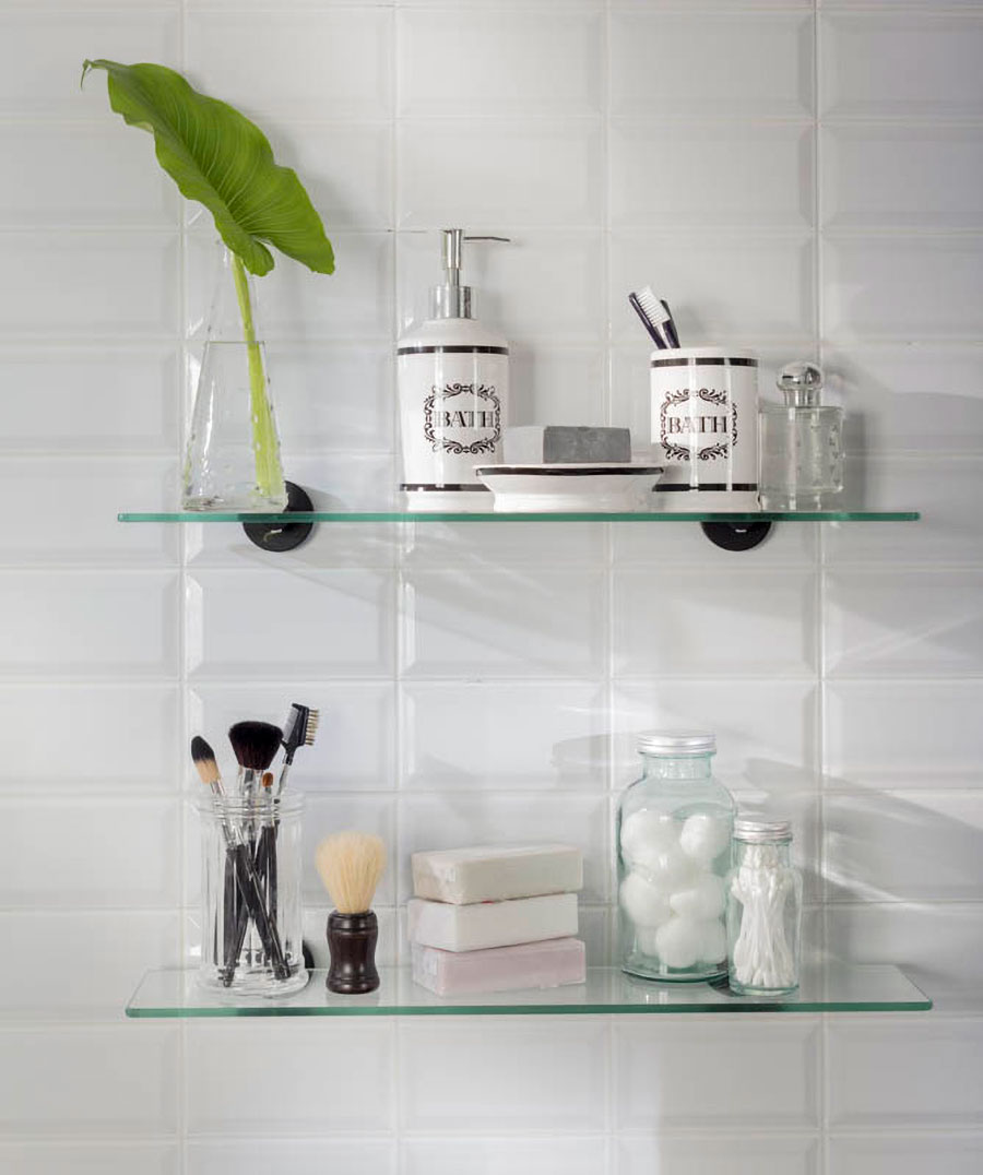 Dos repisas de vidrio para baño, con artículos de aseo, brochas de maquillaje y un jarrón transparente con planta. 