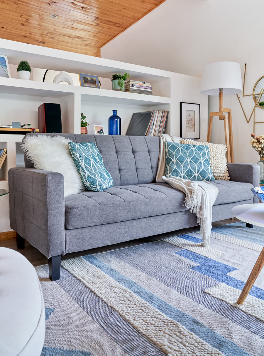 Living con un futón gris con varios cojines de tonos azules, mostazas y blancos y una manta blanca. Está sobre una alfombra de tonos azules y crema y frente a un estante blanco.