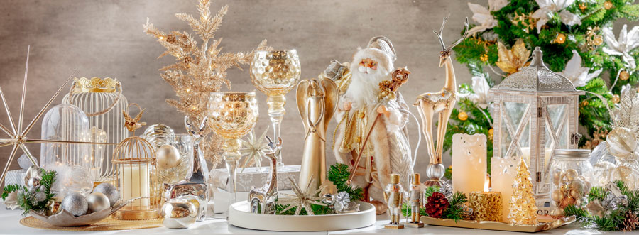 Bodegón de decoración navidad, colección glam. Se ven candelabros de vidrio, bandejas, figuras de Santa y un ciervo, jaulas, estrellas y esferas en tonos dorados y blancos.