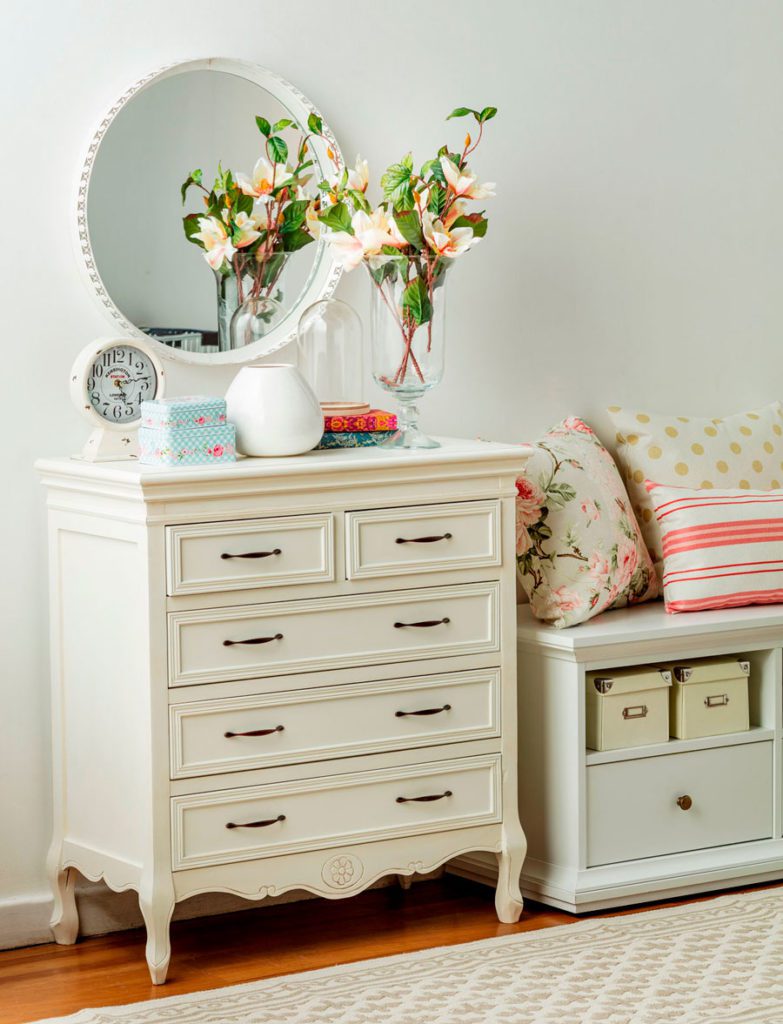 Cajonera blanca de estilo romántico, con un jarrón de flores, y cajas decorativas, junto a un espejo redondo