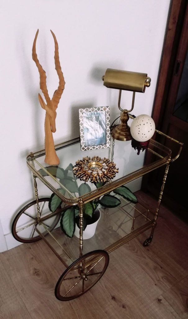 Camarera vintage de bronce y vidrio, decorada con plantas, una lámpara, fotos y una figura animal.