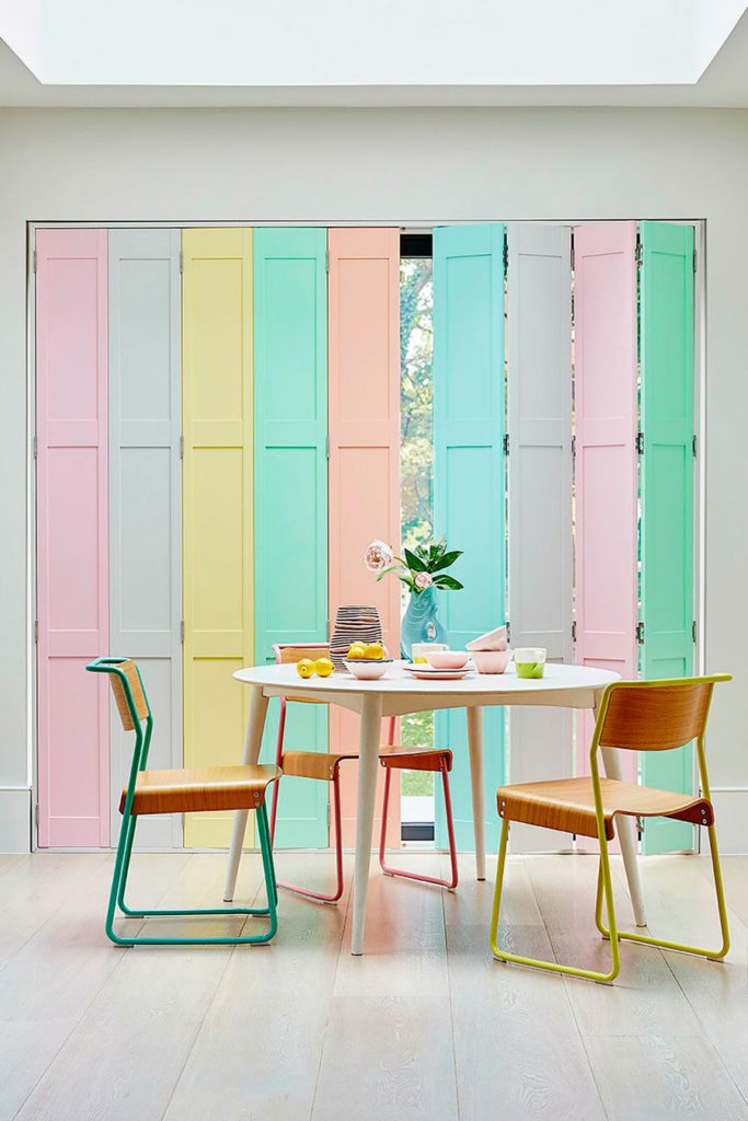 Comedor con sillas de metal y madera pintadas en colores pastel y una ventanal con puertas pintadas en color pastel