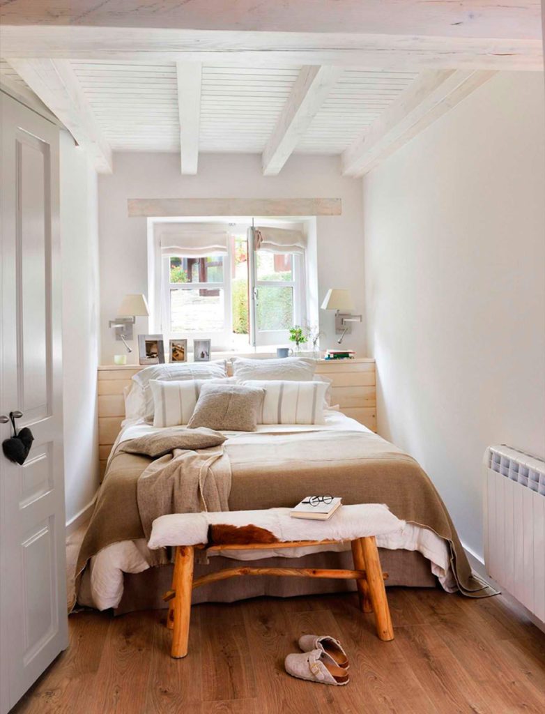 Dormitorio pequeño en tonos blancos y neutros con una banqueta de madera y piel a los pies.