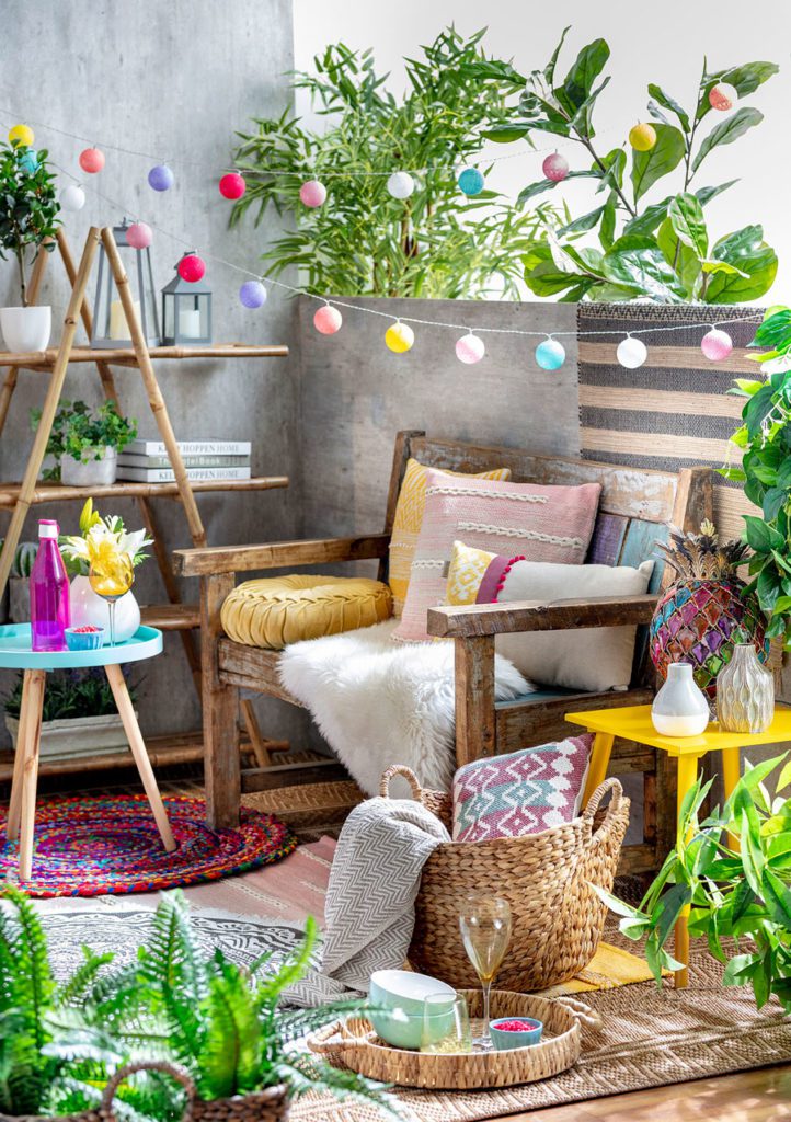 Terraza pequeña con asiento de madera reciclada, decorado con una manta de piel y muchos cojines decorativos de colores, guirnalda de luces multicolor y plantas.
