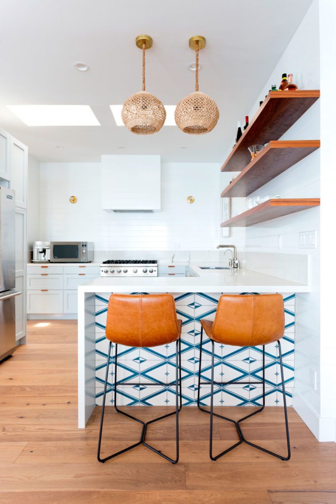Cocina moderna y minimalista, decorada en blanco, con accesorios en tonos madera y azul. Con mesón tipo americano y dos sillas de bar de cuero.
