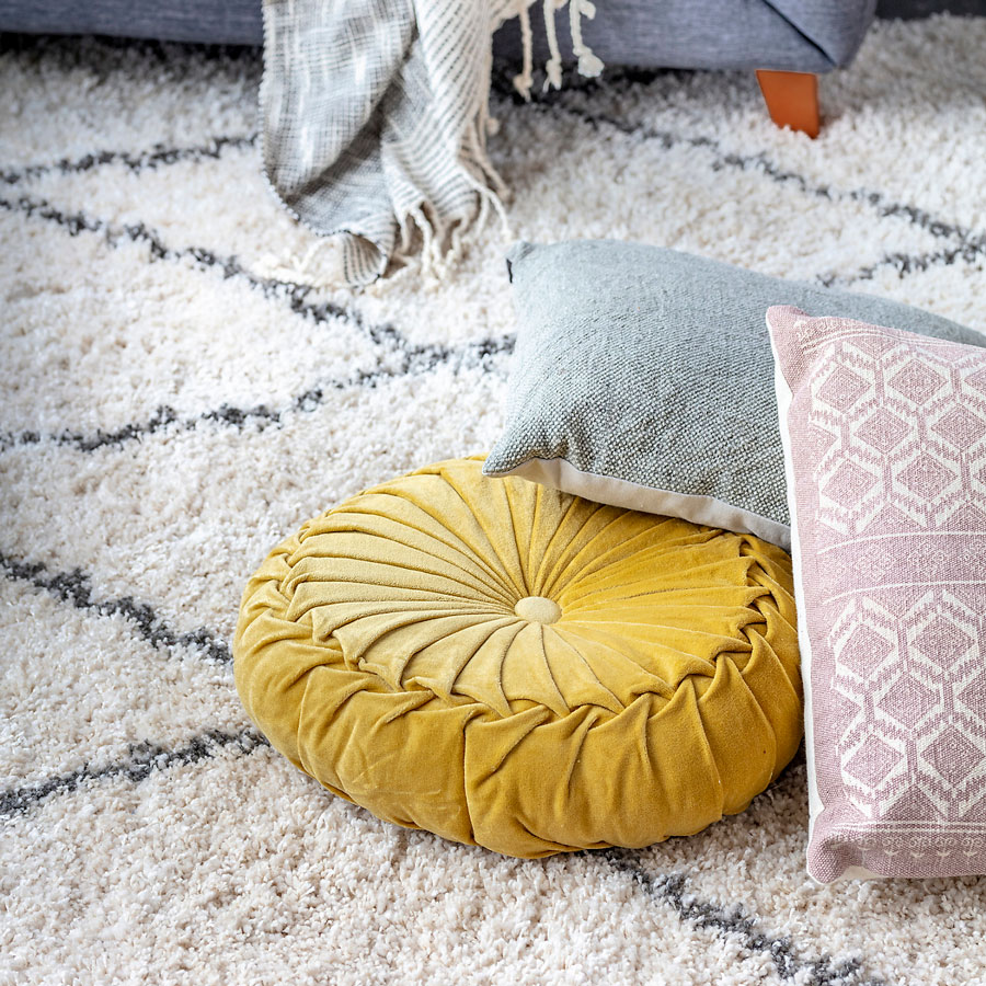 Cojín redondo de terciopelo color mostaza, junto a otros cojines decorativos, sobre alfombra shaggy blanca con motivos geométricos