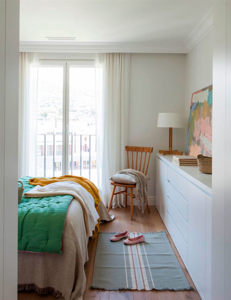 Vista de un dormitorio desde la puerta, se ve la cama con un quilt y una manta en tonos pastel, una silla de madera y un mueble blanco frente a la cama.