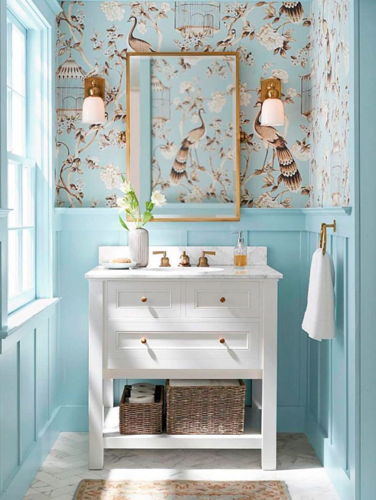 Baño en tonos celeste, con muros mitad madera y mitad papel mural con dibujos de pájaros y jaulas en tonos café claro.