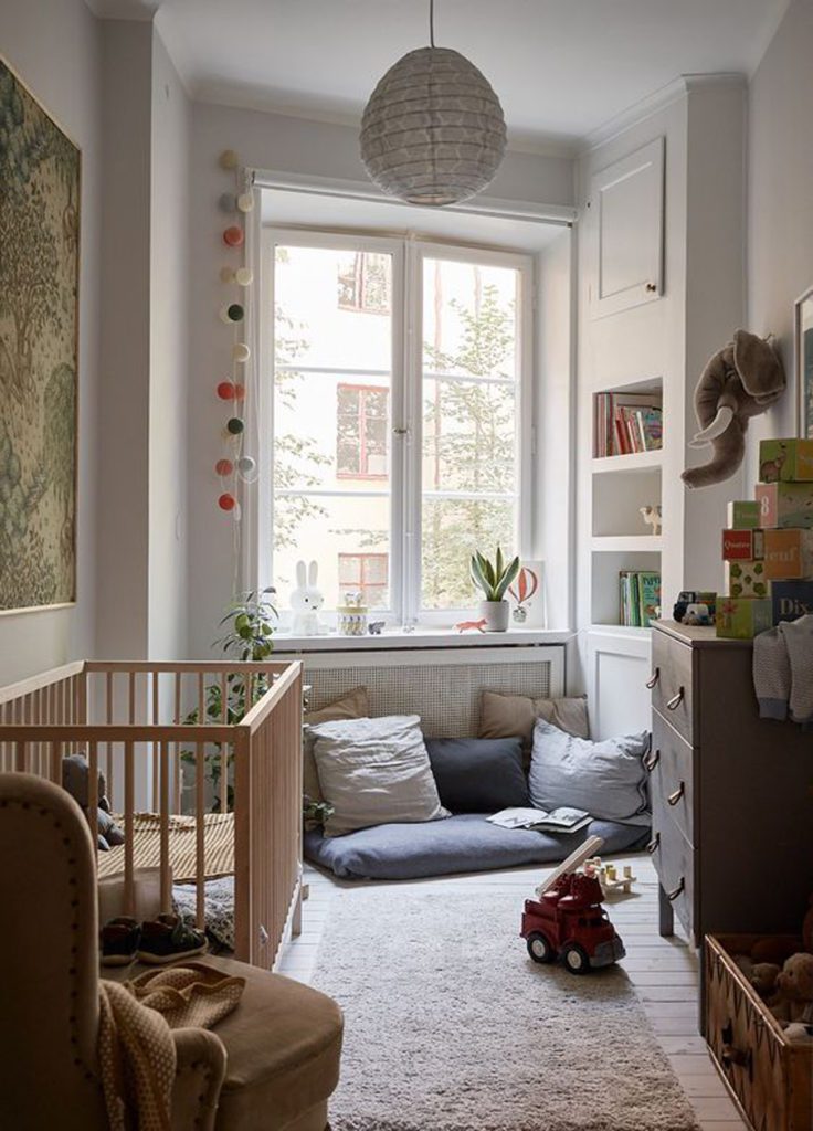 Dormitorio de bebé angosto, con cuna y cajonera de madera, cojines decorativos y alfombra alargada.