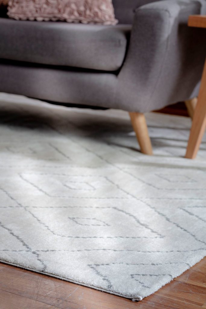 Detalle alfombra en color crudo y diseños geométricos, con un sofá gris con patas de madera