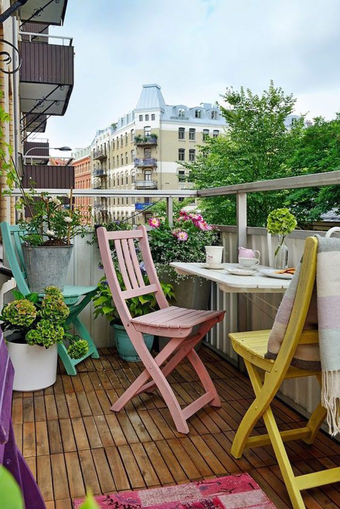 Balcón pequeño, con sillas de madera en colores pastel, una bandeja que sirve de mesa y muchos maceteros con plantas y flores