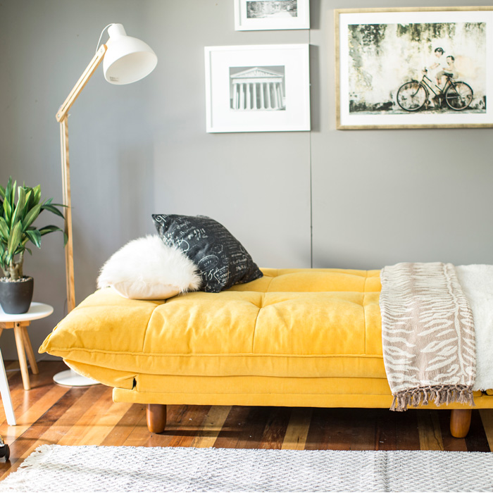 Living con lámpara de pié y futón color amarillo decorado con cojines que combinan con el blanco de las mesas auxiliares, marcos de cuadro y pantalla de lámpara