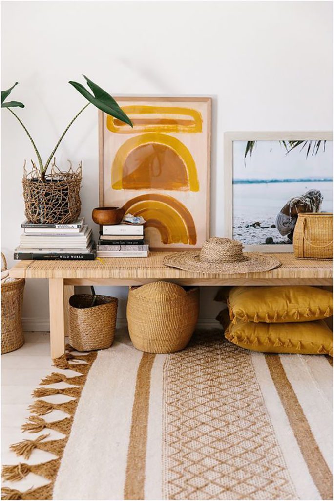 Rincón de una casa con una banqueta de mimbre, acompañada de canastos del mismo material. Toques dorados en cojines y cuadros, además de una planta ornamental de hojas grandes y verdes. Alfombra tejida beige con café. Rincón de una casa con una banqueta de mimbre, acompañada de canastos del mismo material. Toques dorados en cojines y cuadros, además de una planta ornamental de hojas grandes y verdes. Alfombra tejida beige con café.