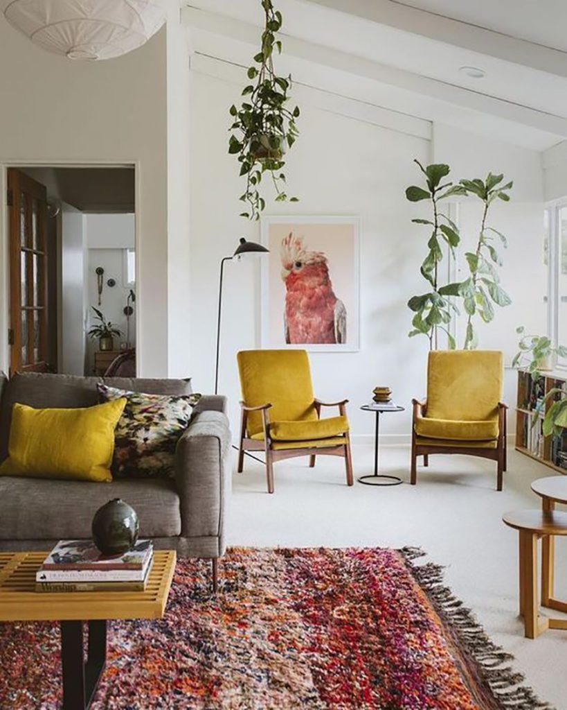 Living y sala de estar con sitiales en tonos mostaza y un sofá color gris con cojines también mostaza. Plantas colgantes y de en maceteros con hojas grandes y verde. Paredes y piso blanco. Living y sala de estar con sitiales en tonos mostaza y un sofá color gris con cojines también mostaza. Plantas colgantes y de en maceteros con hojas grandes y verde. Paredes y piso blanco.