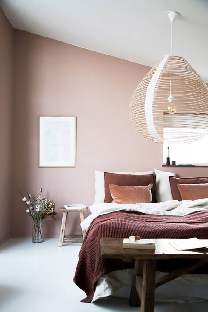 Dormitorio en tonos blush, con cama y banqueta de madera. Cuadros y lámpara colgante con detalles dorados.
