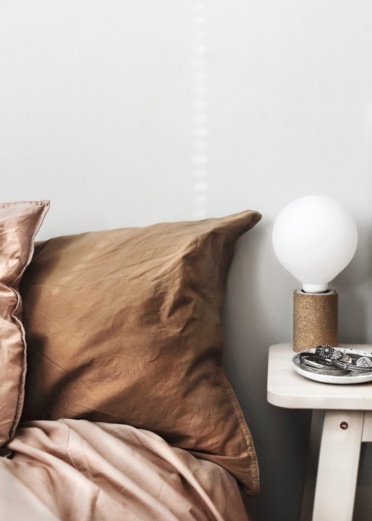 Imagen detalle de una cama con tela de lino, almohada color café y plumón en tono rosa. Velador blanco con una lámpara.